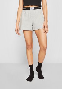 Pantalones cortos de algodón gris con una cinturilla elástica negra que presenta el logo "Calvin Klein". Llevados con calcetines negros hasta el tobillo.
