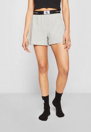 Pantalones cortos de algodón gris con una cinturilla elástica negra que presenta el logo "Calvin Klein". Llevados con calcetines negros hasta el tobillo.
