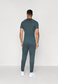 Mężczyzna odwrócony plecami, ubrany w dopasowany ciemnozielony krótki rękaw sportowy T-shirt oraz pasujące dresy, w białych sneakersach na jednolitym tle.