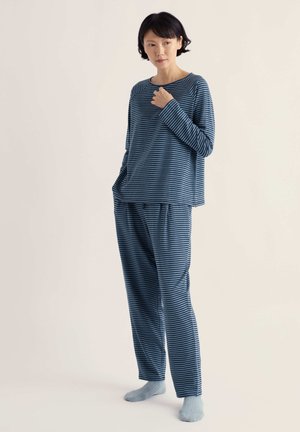Femme debout portant un haut à manches longues rayé bleu et noir et un pantalon assorti avec des chaussettes bleu clair sur un fond uni.