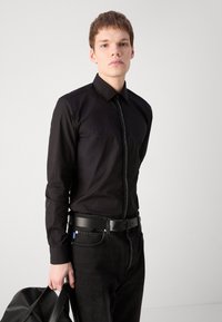 Fekete hosszú ujjú ing, elülső cipzárral és gallérral, fekete farmernadrággal és fekete övvel párosítva. A férfi egy fekete táskát tart.