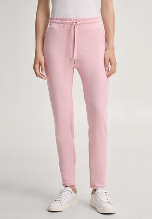 Personne portant un pantalon de survêtement rose clair à cordon et des baskets blanches, debout devant un fond uni.