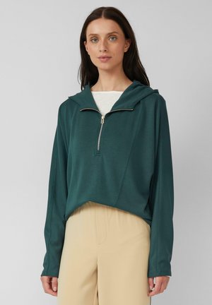 Pullover à capuche teal à manches longues avec une demi-fermeture éclair, présentant une coupe décontractée et une texture douce. Associé à un pantalon beige.