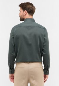 Donkergroene blouse met lange mouwen en een kraag, met knoopmanchetten en een gladde textuur. Draagt in combinatie met beige broek, achterkant zichtbaar.
