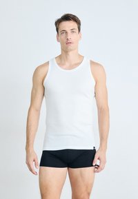 Débardeur blanc en tissu léger, avec un col large et un design sans manches. Le modèle porte un short noir ajusté.