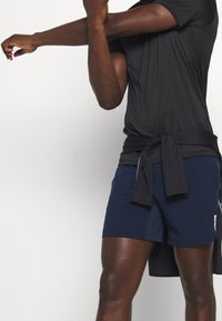 Hombre vestido con camiseta negra y pantalones cortos azul marino estirando su brazo derecho a través del pecho sobre un fondo blanco.