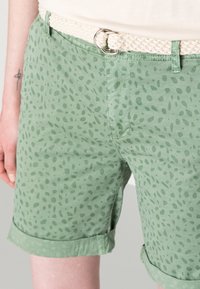 Pantalones cortos verdes con un diseño de patrón de hojas, dobladillo enrollado y un cinturón trenzado blanco con hebilla plateada. Tela de algodón, estilo casual.