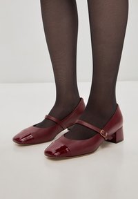Burgundy lakkanahkasa Mary Jane -kengät, joissa on remmi ja neliömäinen varvas, alhaalla oleva loiva korko ja sileä pinta.
