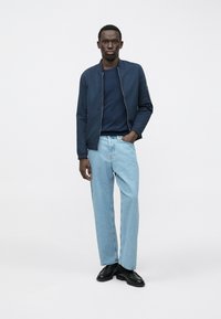 Mann steht in einer marineblauen Bomberjacke, dunklem blauen Hemd, hellblauen Jeans und schwarzen eleganten Schuhen vor einem schlichten weißen Hintergrund.