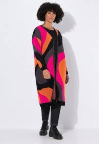 Femme aux cheveux bouclés portant un long cardigan multicolore avec des motifs roses, orange, noirs et gris, un pantalon noir et des bottes noires, debout.