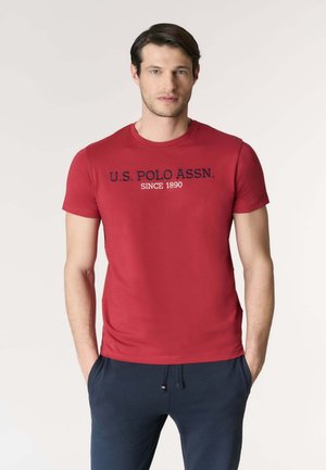 Uomo che indossa una maglietta rossa U.S. Polo Assn. e pantaloni blu navy, in piedi con le mani nelle tasche su uno sfondo neutro.