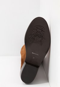 Semelle de bottine en daim marron montrant un blason embossé, le texte "Fabriqué en Italie" et la taille 37 sur un talon noir sur fond blanc.