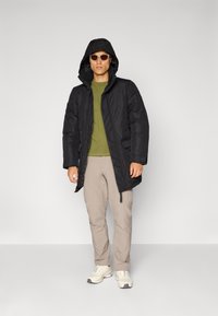 Chaqueta acolchada negra con capucha, llevada sobre una camiseta verde. Pantalones beige y zapatillas ligeras completan el conjunto. Textura oscura y estilo notables.
