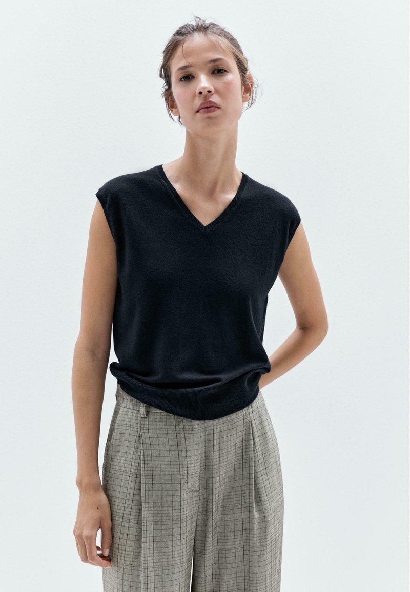 Black sleeveless sweater with a V-neck design, made from a soft material. Worn with gray plaid high-waisted trousers.