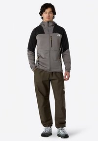 Chaqueta con capucha gris y negra con cierre de cremallera, dos bolsillos delanteros; combinada con pantalones verde oscuro y zapatos de senderismo grises.