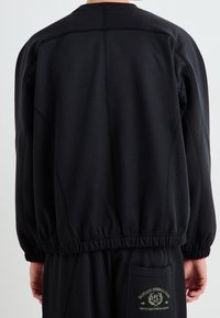 Czarna bluza z luźnym krojem, wyposażona w elastyczne mankiety i dół oraz widoczne logo wyszywane na plecach. Gładka faktura tkaniny.