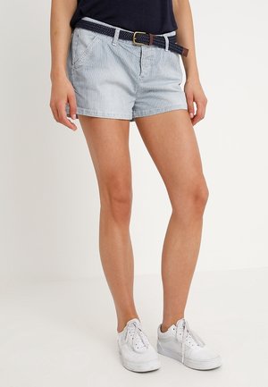 Kvinde iført lyseblå, stribede shorts med navy vævet bælte, navy top og hvide sneakers, stående mod en ensfarvet baggrund.