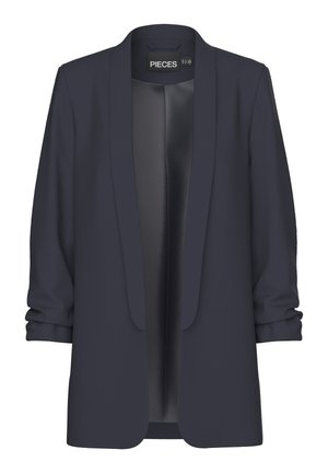 Femme portant un blazer bleu marine aux manches retroussées sur un top noir et un jean bleu taille haute devant un fond uni.