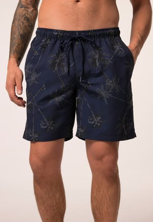 Mężczyzna noszący granatowe szorty kąpielowe z czarnym nadrukiem palm i ściągaczem w pasie, widoczny tatuaż na lewym przedramieniu.