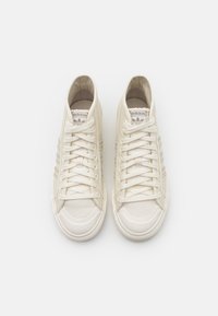 Bílé plátěné tenisky s gumovými podrážkami, které mají tři vodorovné pruhy, kulatou špičku a šněrování. Jemné logo na jazyku.