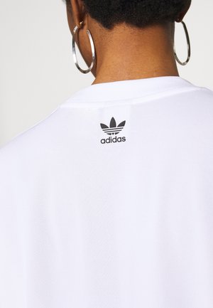 Personne aux cheveux courts et bouclés, portant de grandes boucles d'oreilles argentées et un t-shirt blanc Adidas avec le logo au niveau du haut du dos.