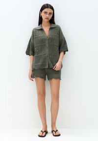Grön stickad cardigan med krage och knappar, med mönster i öppen arbetsteknik. I hop med matchande shorts som har vågiga kanter.