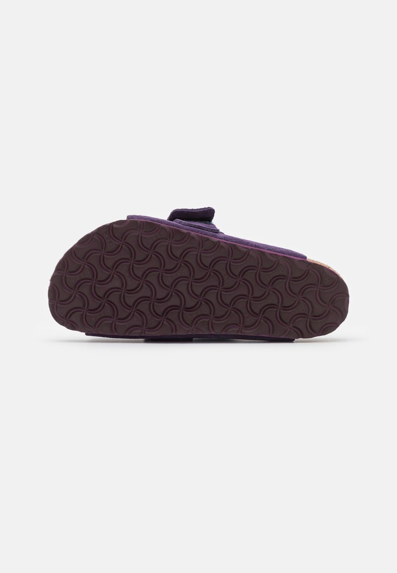Lila Hausschuh mit einer strukturierten Gummisohle, ausgestattet mit einem breiten Riemen und einem Obermaterial aus weichem Stoff. Die Sohle hat ein wellenförmiges Muster für besseren Halt.