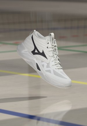 Chaussure de sport blanche avec un logo noir flottant au-dessus d’un sol de salle de sport marqué de lignes colorées de terrain. Aucune personne visible.
