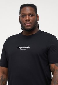 Sort bomulds T-shirt med korte ærmer, der har en hvid logo tekst "Originals Studio" på brystet. Regular fit, glat tekstur.