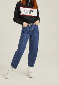 Blå denimjeans med hög midja och lös passform, parat med en svart och vit zip-up sweatshirt med en röd rand och logotyp. Vita sneakers.