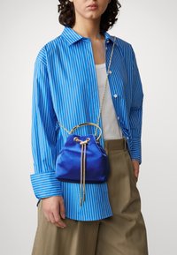 Piccola borsa a secchiello blu in satin con tracolla in oro, dotata di chiusura a coulisse e un accento circolare. Indossata con una camicia a righe blu.