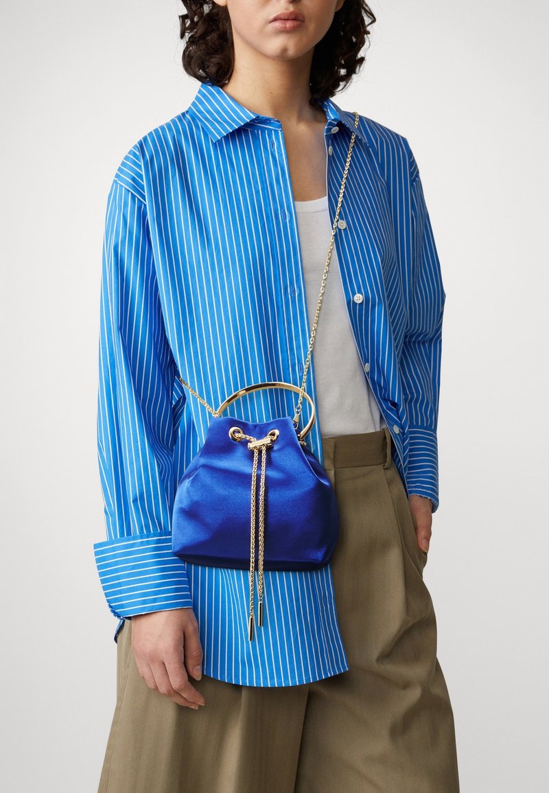 Piccola borsa a secchiello blu in satin con tracolla in oro, dotata di chiusura a coulisse e un accento circolare. Indossata con una camicia a righe blu.