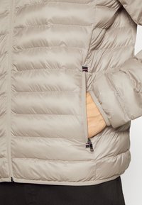 Giacca puffer beige chiaro con design trapuntato, tasca laterale con zip e polsini elasticizzati. Presenta una texture di tessuto liscia e lucente.