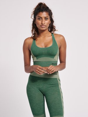 Frau mit lockigem Haar in einem grünen Sport-BH und passenden Leggings, die vor einem schlichten hellen Hintergrund steht und mit verschränkten Händen nach vorne schaut.