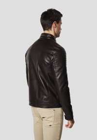 Must nahast jope, kärbitud lõikega, millel on polsterdatud õlad ja kõrge krae, kombineeritud heledate pükstega.