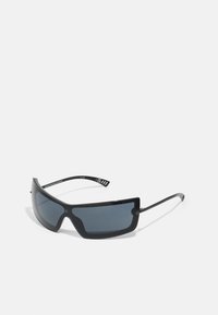 Des lunettes de soleil noires avec des verres angulaires et larges et des branches élégantes. Les verres sont teintés de gris, et la monture présente une finition mate avec des accents métalliques.