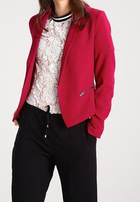 Fuchsia blazer med en strukturerad design och dragkedjefickor, bärs över en vit spetsblus, i kombination med svarta, dragsnöringsbyxor.