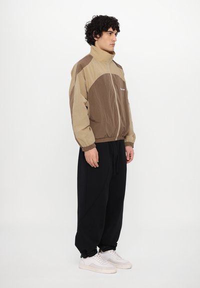 Jachetă windbreaker în nuanțe de maro și bej, cu guler înalt, închidere cu fermoar și material texturat. Asortată cu pantaloni negri, largi și adidași albi.