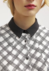 Kobieta w czarno-białą w kratkę bluzce z solidnym czarnym kołnierzykiem w szpic i czarnymi guzikami.