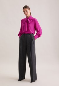 Blusa fucsia con un grande fiocco, maniche lunghe e polsini con bottoni; abbinata a pantaloni ampi a vita alta grigio scuro con tasche.