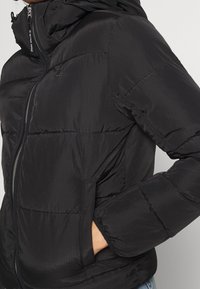 Veste matelassée noire avec un devant zippé, texture matelassée, col haut et capuche avec cordon de serrage. Le matériau semble brillant et lisse.