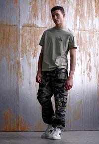 T-shirt en coton olive avec logo, associée à un pantalon cargo camouflage doté de multiples poches, et des baskets blanches avec des accents bleus.