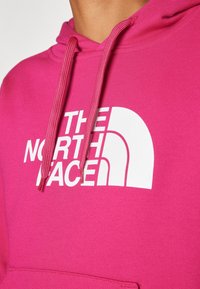 Sudadera con capucha color rosa fuerte hecha de tela suave, con un gran logo blanco "THE NORTH FACE" en el frente, con cordones ajustables y un bolsillo tipo canguro.