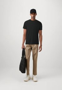 Černé tričko, béžové kalhoty, černá prošívaná taška, bílé tenisky a černá čepice. Model stojí uprostřed na neutrálním pozadí.