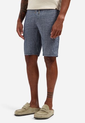 Man draagt blauwe, gestructureerde korte broek en beige instaploafers, staand met ontspannen armen langs het lichaam tegen een lichte achtergrond.