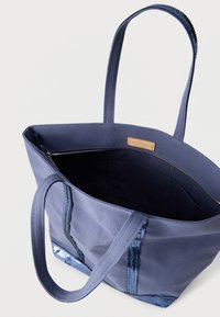 Sac fourre-tout en toile bleue avec double poignée et bordure à sequins, montrant l'intérieur avec une poche zippée et une petite étiquette de marque en cuir.