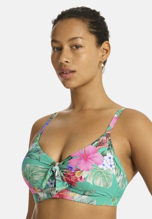 Kobieta z ciemnymi włosami związanymi do tyłu, ubrana w zielony kwiecisty top bikini z różowymi i białymi kwiatami na jednolitym tle.