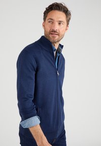 Marineblauer Reißverschluss-Pullover mit hohem Kragen, gerippten Bündchen und Taschen. Über einem hellblauen Hemd getragen. Weiche Textur, lässiges Design.