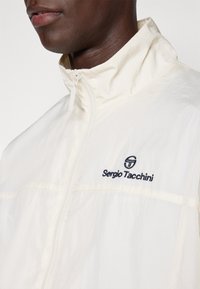 Hvit zip-up jakke med høy krage, med en svart brodert logo på brystet, glatt tekstur og subtil glans.