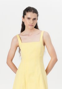 Vestido amarillo de cuello cuadrado con tirantes anchos, diseño ajustado y tela texturizada, que llega a la mitad de la pantorrilla. Silueta simple y elegante.